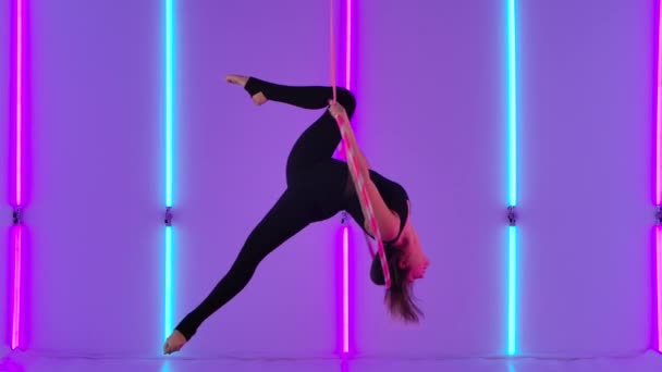 Urheilullinen antenni tyttö akrobaatti musta trikoo suorittaa korkeudella ja suorittaa akrobaattisia elementtejä antenni vanne. Nuoren taiteilijan sirkusesitys kirkkaiden neonvalojen keskellä. Hidastus. — kuvapankkivideo