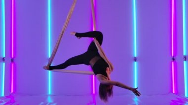 Hava ipeğinde genç bir sirk sanatçısının akrobatik performansı. Siyah taytlı bir kız parlak neon ışıkların arka planına karşı havada fırıl fırıl döner. Yavaş çekim.