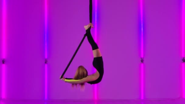 Flexibilní mladý gymnastka visí hlavou dolů na vzduchovém koši a provádí akrobatické prvky. Dívka v černém trikotu hraje cirkusovou show na pozadí jasných neonových světel. Zpomalený pohyb. — Stock video