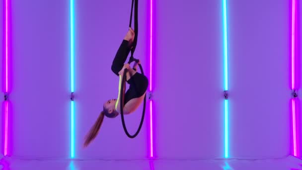 La gimnasta joven y flexible cuelga boca abajo en un aro de aire y realiza elementos acrobáticos. Chica en maillot negro realiza espectáculo de circo contra el telón de fondo de luces de neón brillantes. Movimiento lento. — Vídeos de Stock