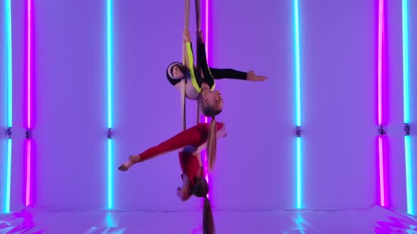 Dos gimnastas aéreos realizan rotación y torzal en seda aérea. Chicas jóvenes en negro y rojo maillot equilibra contra el fondo de luces de neón brillantes en el estudio. Movimiento lento. — Vídeos de Stock