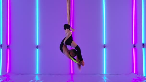 Equilibrio gimnasta estirada y girando sobre seda aireada. Chica joven en maillot negro realiza elementos acrobáticos en el estudio contra el fondo de luces de neón brillantes. Movimiento lento. — Vídeo de stock