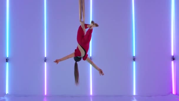 Aerial gymnast performing aerial acrobatic dance on silks in the studio. A young girl in a red leotard demonstrates stretching against a background of bright neon lights. Slow motion. — Stock Video