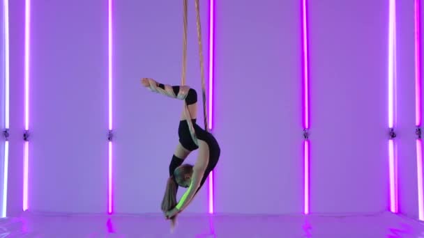 Giovane ragazza equilibrista in body nero ruota su seta ariosa e dimostra stretching. Difficili acrobazie acrobatiche in altezza sullo sfondo di luci al neon luminose. Rallentatore. — Video Stock