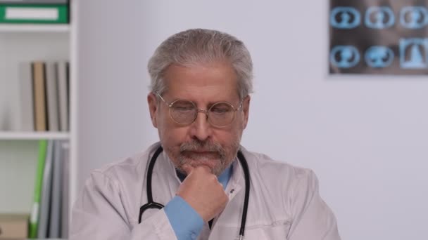 Aged male doctor in white medical coat with concentration thinks. Doc with gray hair and glasses sits at a table in a hospital office. Close up. Slow motion. — Vídeo de stock