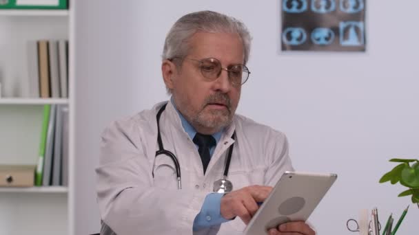 Aged male doctor in white medical coat examines the patient medical history on a tablet and makes notes. Doc with gray hair and glasses sits at a table in a hospital office. Close up. Slow motion. — Stockvideo