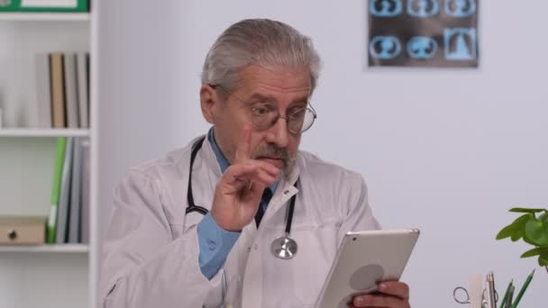 Лікар у віці чоловіків у білому медичному пальто робить конференц-дзвінок на планшет, консультуючи дистанційного пацієнта онлайн у відео чаті. Лікар з сірим волоссям сидить за столом у лікарняному кабінеті. Близько. Повільний рух . — стокове відео