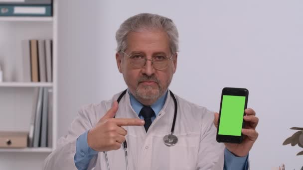 Aged male doctor holds smartphone with green screen chroma key, points at it with finger and makes thumb up gesture. Doc in white medical coat sits at table in hospital office. Close up. Slow motion. — Stockvideo