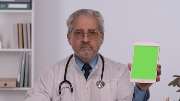 Aged male doctor holds tablet with green screen chroma key, points at it with finger and makes thumb up gesture. Doc in white medical coat sits at table in hospital office. Close up. Slow motion. — Stock Video