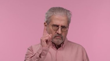 Portrait of an elderly man with glasses threatening shaking her index finger. Gray haired pensioner grandfather with beard wearing shirt posing on pink studio background. Close up. Slow motion.