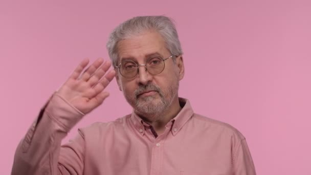 Porträt eines älteren Mannes mit Brille, winkender Hand und Geste. Der grauhaarige Rentner-Opa mit Bart trägt ein Hemd, das auf rosa Studiohintergrund posiert. Nahaufnahme. Zeitlupe. — Stockvideo