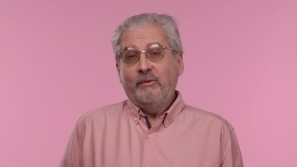 Portrait of an elderly man with glasses is reporting and telling lot of interesting information. Gray haired pensioner grandfather with beard posing on pink studio background. Close up. Slow motion. — Stock Video