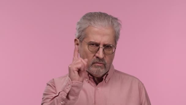 Portrait of an elderly man with glasses threatening shaking her index finger. Gray haired pensioner grandfather with beard wearing shirt posing on pink studio background. Close up. Slow motion. — ストック動画
