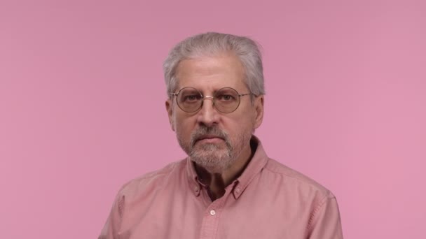 Portrait of an elderly man looks at camera with excitement, then sighs in relief, received pleasant information. Gray haired pensioner posing on pink studio background. Close up. Slow motion. — Video