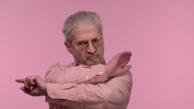 Portrait of an elderly man with glasses strictly gesturing with hands shape meaning denial saying NO. Gray haired pensioner grandfather posing on pink studio background. Close up. Slow motion.