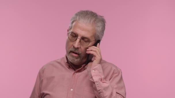 Porträt eines älteren Mannes mit Brille, der für sein Handy spricht. Der grauhaarige Rentner-Opa mit Bart trägt ein Hemd, das auf rosa Studiohintergrund posiert. Nahaufnahme. Zeitlupe. — Stockvideo