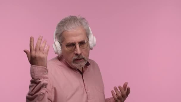 Portrait of an elderly man with glasses dancing and enjoying music in big white headphones. Gray haired pensioner grandfather with beard posing on pink studio background. Close up. Slow motion. — Stock Video