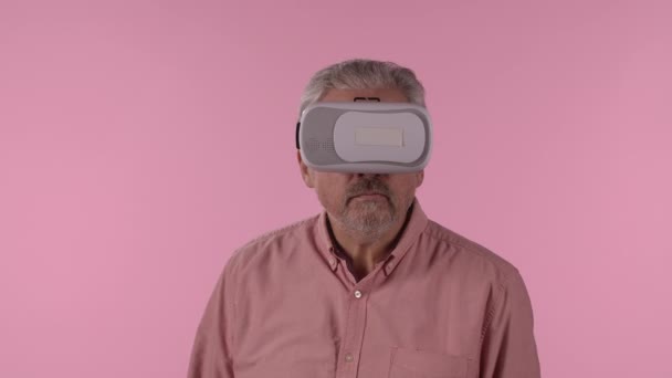 Portrait of an elderly man with virtual reality headset or 3d glasses. Gray haired pensioner grandfather with beard wearing shirt posing on pink studio background. Close up. Slow motion. — 비디오
