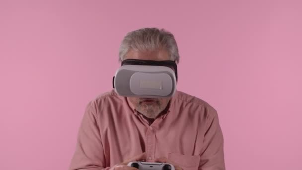 Portrait of an elderly man with virtual reality headset playing video game using wireless controller with joy in victory. Gray haired pensioner posing on pink studio background. Close up. Slow motion. — Vídeos de Stock