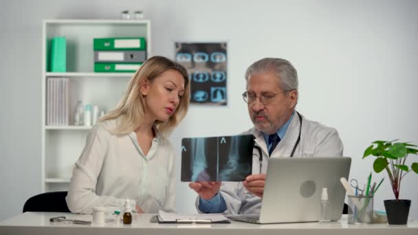 Senior doctor consults patient by showing her picture of leg and discusses treatment of disease with her. Patient listens doc in white medical suit sitting at table in hospital office. Slow motion. — Vídeo de stock