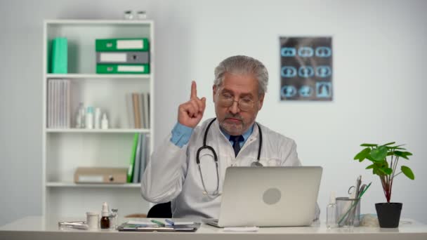 Пожилой врач в белом медицинском халате, печатающий на ноутбуке, думая пристально, появляется идея и продолжает печатать. Док сидит за столом в больничном кабинете. замедленное движение. — стоковое видео