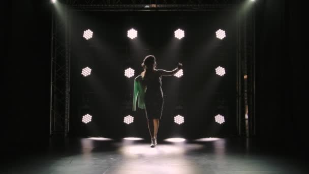 Een slanke brunette van middelbare leeftijd loopt in een donkere studio tegen een achtergrond van felle lichten. Een vrouw in een korte jurk en witte loafers loopt en kijkt intens naar beneden. Silhouet. Langzame beweging. — Stockvideo