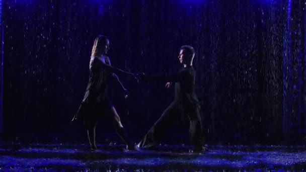 Tancerze towarzyscy tańczący namiętny latynoamerykański taniec bachaty w ulewnym deszczu. Sylwetki młodej pary w mokrych ubraniach na czarnym tle studia z kroplami wody i niebieskim światłem. Zwolniony ruch. — Wideo stockowe
