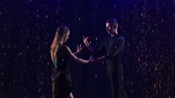 Bachata apaixonado dança entre as gotas de chuva realizadas por um jovem casal sensual de dançarinos. Os parceiros se movem em uma dança em um fundo de estúdio preto entre as luzes de néon azul. Fecha. Movimento lento. — Vídeo de Stock