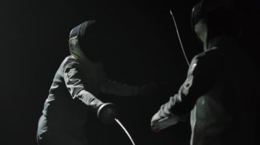 İki profesyonel eskrimci folyo eskrim ustalığını gösteriyor. Beyaz üniformalı ve koruyucu maskeli atletlerin siluetleri siyah stüdyo arka planında düello yapıyor. Ağır çekim. Kapat..