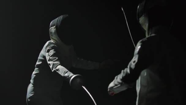 Two professional fencers demonstrate their mastery of foil fencing. Silhouettes of athletes in white uniforms and protective masks fighting duel on black studio background. Slow motion. Close up. — Stock Video