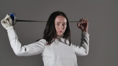 Beyaz üniformalı genç bir eskrimci kadın portresi elinde bir rapier ile gri bir stüdyo arka planında poz veriyor. Eskrim okulu posteri. Ağır çekim. Kapat..