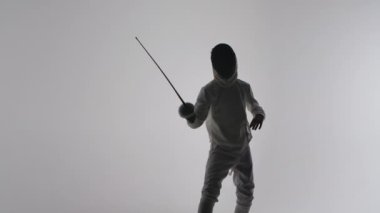 Beyaz üniformalı ve maskeli genç bir çocuğun silueti gri stüdyo arka planında Rapier 'ı ustaca kullanıyor. Saldırı ve savunma eğitimi, spor okulunda eskrim dersi. Ağır çekim. Kapat..