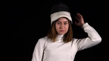 Kendine güvenen bir eskrimcinin portresi kameraya bakıyor ve siyah bir stüdyonun arka planına koruyucu bir maske takıyor. Beyaz üniformalı kadın sporcu elinde iple poz veriyor. Kapat..