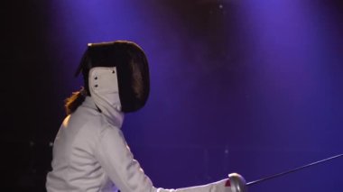 İki bayan sporcu, folyo becerilerini eskrim turnuvasında geliştiriyor. Genç kadın eskrimciler, koyu dumanlı bir stüdyoda mavi ışıkla düello yapıyorlar. Kapat..