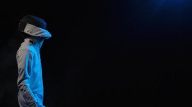 Genç silahşör koruyucu maske takıyor. Beyaz üniformalı bir sporcu elinde rapier ile siyah dumanlı arka planda mavi ışıkla poz veriyor. Ağır çekim. Kapat..