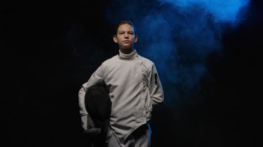 Kılıç ustasının portresi koruyucu maske kaskı takıyor. Beyaz üniformalı bir sporcu elinde rapier ile siyah dumanlı arka planda mavi ışıkla poz veriyor. Ağır çekim. Kapat..