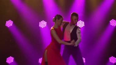 Parlak mor ışıklarla karanlık bir stüdyoda Arjantin tangosuyla dans eden tutkulu genç çift. Kırmızı elbiseli kadın ve şık takım elbiseli adam tatilde dans gösterisi yapıyorlar. Siluetler. Kapat..