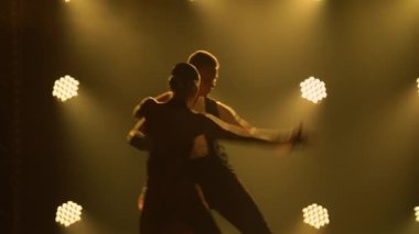 Arjantin dansının elementlerini sergileyen bir çift dansçının siluetleri karanlık stüdyonun dumanlı sarı arka planına karşı ortaya çıkıyor. Kapat..