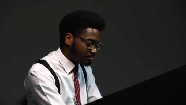 Handsome African American man in a stylish suit and glasses sings and composes himself on the grand piano with pleasure. A black performer performs on a gray background in the studio. Close up. — Stock Video