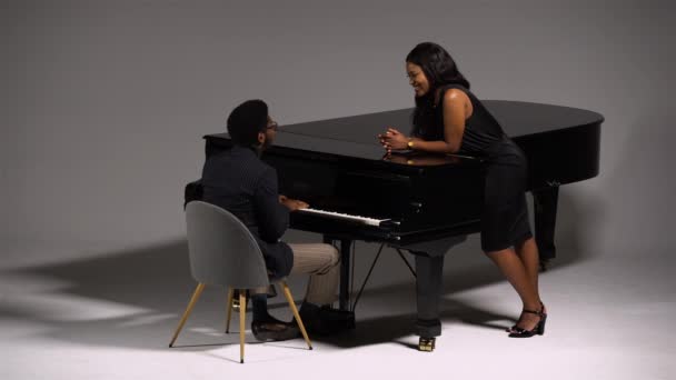 Elegante afroamericano canta románticamente y toca una melodía en el piano de cola para su amada. Una joven escucha y disfruta de la melodía. Un par de negros sobre fondo gris. — Vídeos de Stock