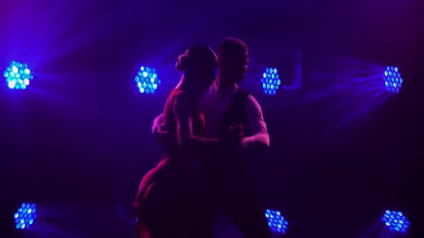 Пара танго-исполнителей танцуют вместе, страстно кружась и держась за руки в темной студии с яркими голубыми огнями. Силуэты стройных фигур. Закрыть. — стоковое видео