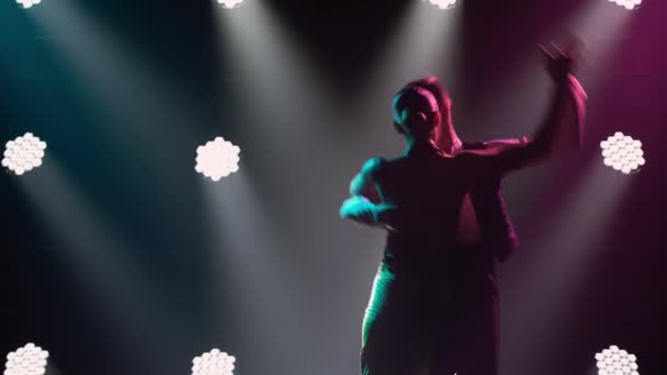 A couple is dancing a fast rhythmic tango dance in a dark studio with bright spotlights. A passionate woman in a red dress and a man in a suit are dancing holding hands. Silhouettes. Close up. — Stock Video