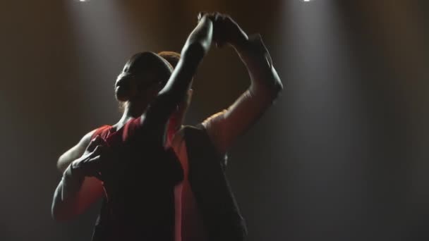 Uma dança de tango apaixonada executada por um par de dançarinos de salão. Homem e mulher elegantes dançando elementos de dança latina em um estúdio escuro com luzes brilhantes. Silhuetas. Fecha. Movimento lento. — Vídeo de Stock