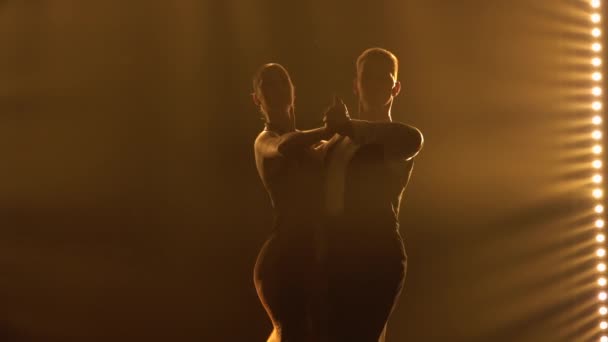 Sylwetki para tancerzy wykonujących elementy argentyńskiego tańca pojawiają się na dymiącym żółtym tle ciemnego studia. Dance show w wykonaniu namiętnych partnerów tanecznych. Zamknij drzwi. Zwolniony ruch. — Wideo stockowe