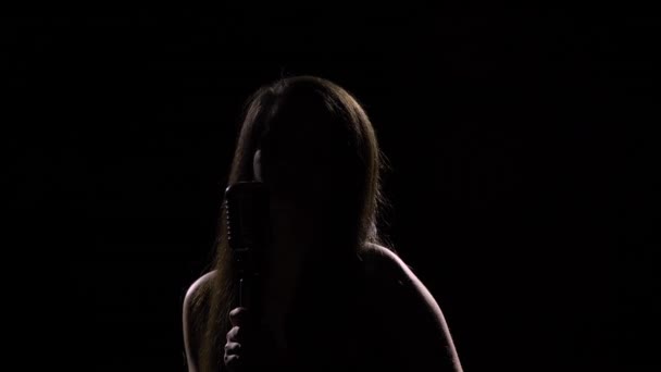 Силуэт молодой женщины, поющей в ретро-микрофон. Женщина-певица с длинными волосами танцует под ритм мелодии и касается микрофона руками на фоне черной студии. Закрыть. — стоковое видео