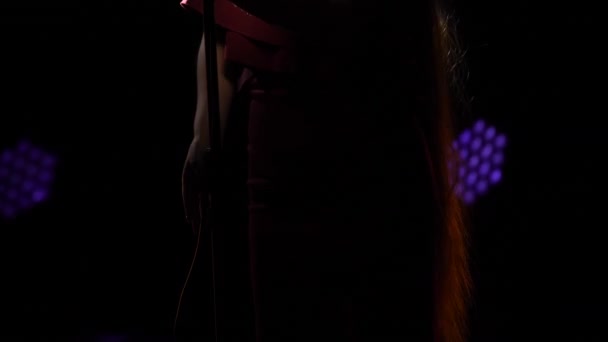 Gyönyörű nő hosszú haj piros ruhában áll a színpadon, és énekel a vintage mikrofon. Singer szexuálisan mozog a sötét stúdióban a kék fények árnyékában. Női sziluett közelről. — Stock videók