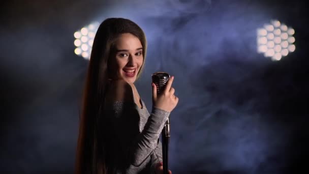 Hermosa joven sexy mujer con el pelo largo en un vestido de noche canta en un micrófono vintage. La cantante sonríe y se mueve elegantemente sobre un fondo ahumado en un estudio oscuro. De cerca.. — Vídeo de stock