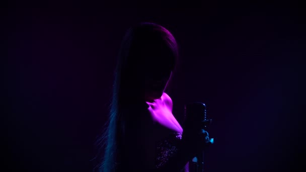 Attraente cantante donna che esegue musica dal vivo in microfono retrò alla festa. Performance di un artista dai capelli lunghi illuminato da luci blu e viola in studio scuro. Silhouette. Da vicino.. — Video Stock