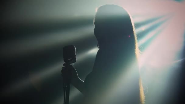 La música femenina con un micrófono retro profesional actúa en el escenario en la oscuridad en el centro de atención. Una mujer sexy en un vestido brillante abierto canta una canción alegre rápido. Silueta. De cerca.. — Vídeos de Stock