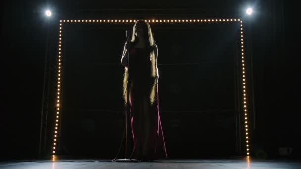 Bottom view of a chic female soloist in a long red dress singing on an empty stage into a retro microphone. Artisca with long hair gives a concert at a music festival in the dark. Slow motion. — Stock Video